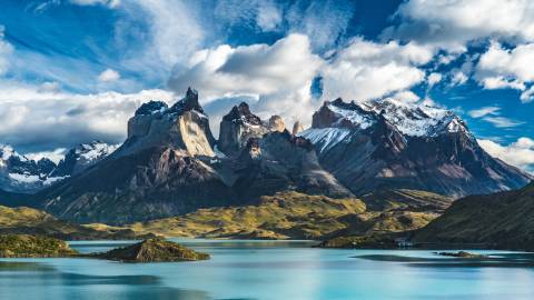 Majestic snow-capped mountains rise against a dramatic sky, reflected in the serene turquoise waters of a lake in the foreground, creating a breathtaking natural landscape.