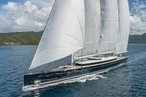 A majestic dark blue sailing yacht glides through the blue ocean with sails billowing under a cloudy sky.