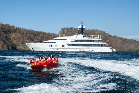 A family enjoys a thrilling ride on a tube towed behind a luxury yacht near a tropical island.