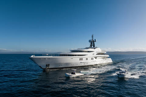 A sleek white superyacht cruises through the deep blue ocean, accompanied by two smaller boats.
