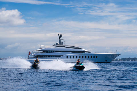 Two jet skiers race across the waves in front of a luxurious superyacht under a cloudy blue sky.