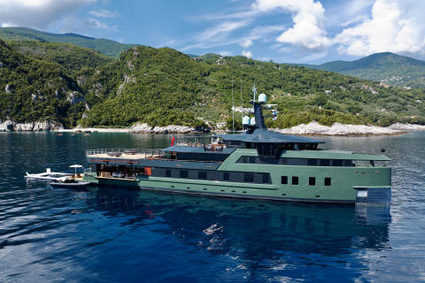 A sleek green yacht named Pink Shadow cruises on calm blue waters near a lush, mountainous coastline.