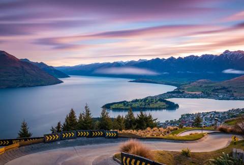 A winding luge track overlooks a serene lake and snow-capped mountains at sunset.