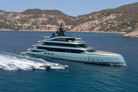 A luxurious turquoise yacht cruises through the azure waters, accompanied by jet skis and a tender.