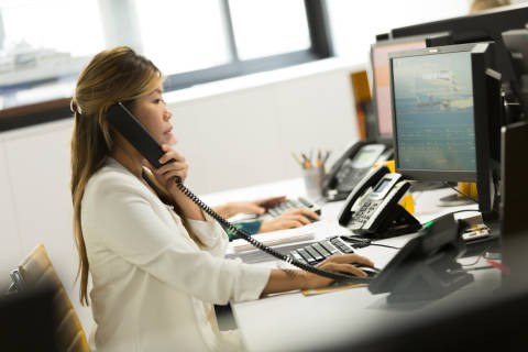 A woman in a white blouse is sitting at a desk, working on a computer and talking on a landline phone in an office setting with various office equipment and a window in the background.