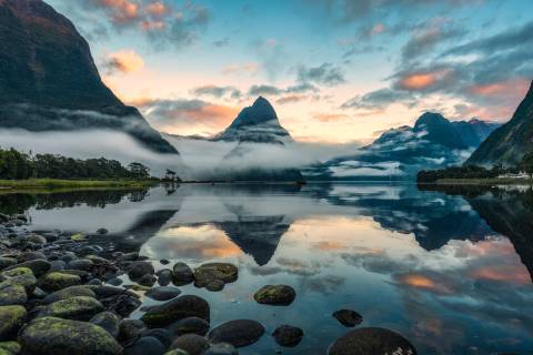 Misty mountains rise from a tranquil lake reflecting the soft hues of a sunrise.