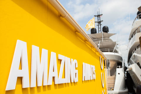 White letters spelling "AMAZING AWAITS" are mounted on a bright yellow wall, with yachts docked in the background under a cloudy sky.