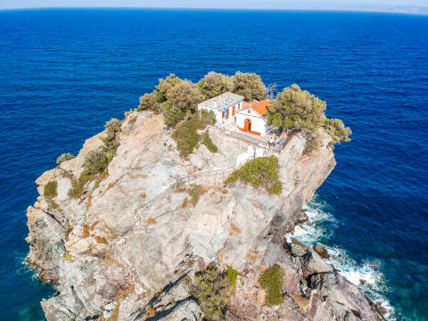 A picturesque white chapel with an orange roof perches atop a rocky outcrop surrounded by the deep blue Aegean Sea.