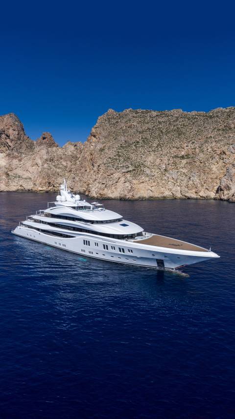 A magnificent white superyacht glides through deep blue waters near a rugged, rocky coastline under a clear sky.