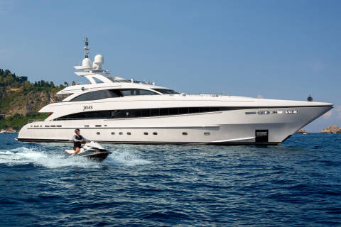 A person rides a jet ski through choppy blue water in front of a large white yacht under a clear sky.