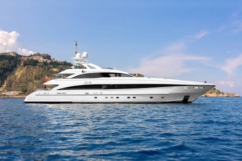 A luxurious white yacht glides across the deep blue sea under a clear sky, with a picturesque coastal town nestled on the hills in the background.
