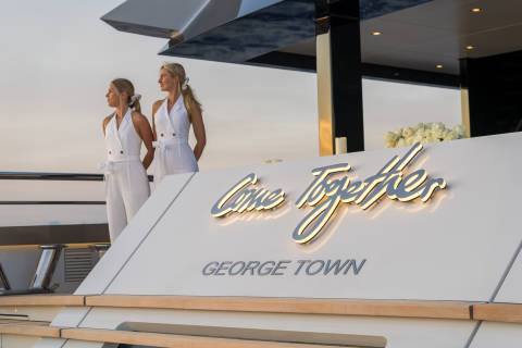 Two elegantly dressed women stand on a yacht deck with a lit "Come Together" sign and a bouquet of roses.