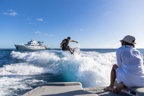 A man in mid-air performs a trick on a wakeboard, leaving a trail of spray behind him as a yacht sails in the distance.