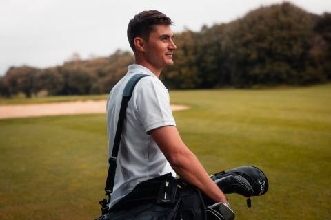 A young man in a white shirt stands on a grassy field, with a golf bag slung over his shoulder, surrounded by a forested landscape in the background.