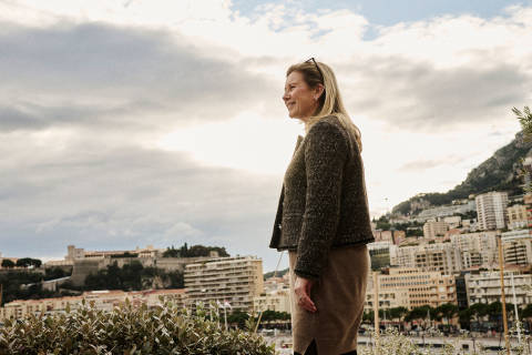 A woman in a warm sweater stands in the foreground, with a cityscape of high-rise buildings and a cloudy sky visible in the background.