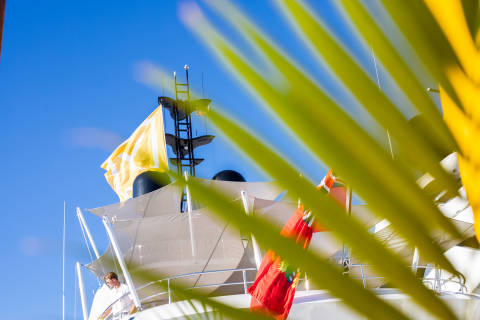 A man in a white shirt looks out from the deck of a yacht flying colorful flags under a bright blue sky.