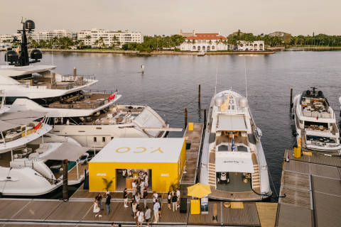Luxury yachts are docked along a waterfront promenade, showcasing opulent vessels and a vibrant outdoor event.