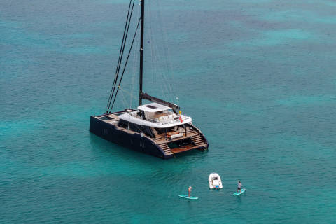 A large catamaran sailboat floats on the serene, turquoise waters, with two people paddleboarding nearby.