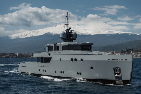 A large, sleek white yacht navigates the calm waters, with snow-capped mountains visible in the background against a cloudy sky.