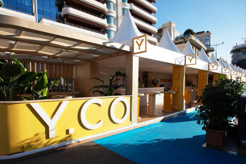 In the foreground, a bright yellow structure with "Y.CO" emblazoned on it is set against a vibrant blue carpet, while the background features a row of white tents with yellow accents and modern buildings under a clear sky.