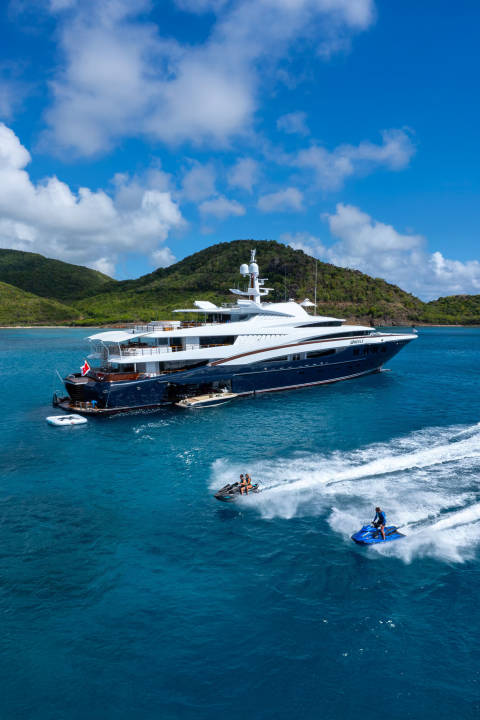 A large blue and white yacht floats on turquoise water in the foreground, with two jet skis zipping by, while lush green hills under a bright blue sky with fluffy clouds form the background.