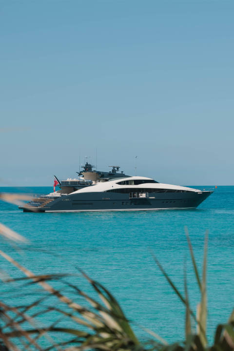 A sleek, dark yacht cruises on turquoise water, with blurred green foliage in the foreground and a clear blue sky in the background.