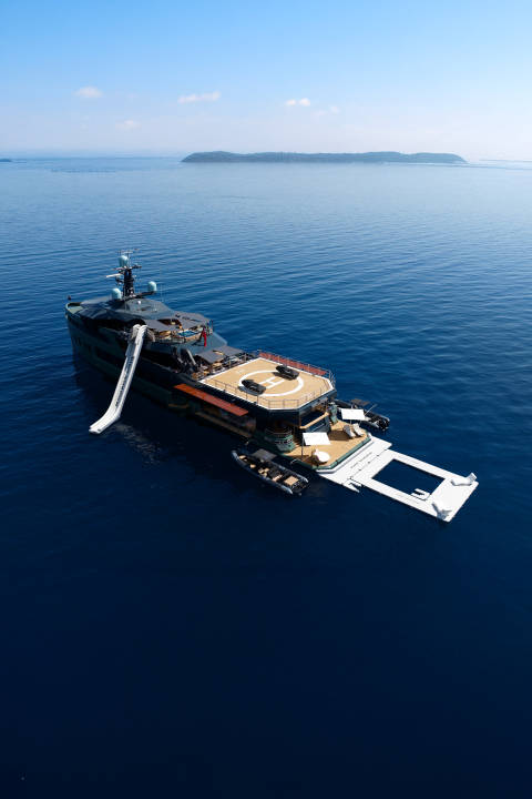 A large yacht with a waterslide and helipad floats on the deep blue ocean, with a distant, tree-covered island on the horizon.