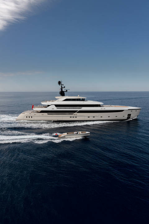 A large white yacht cruises through dark blue water, trailed by a smaller boat, under a clear blue sky.