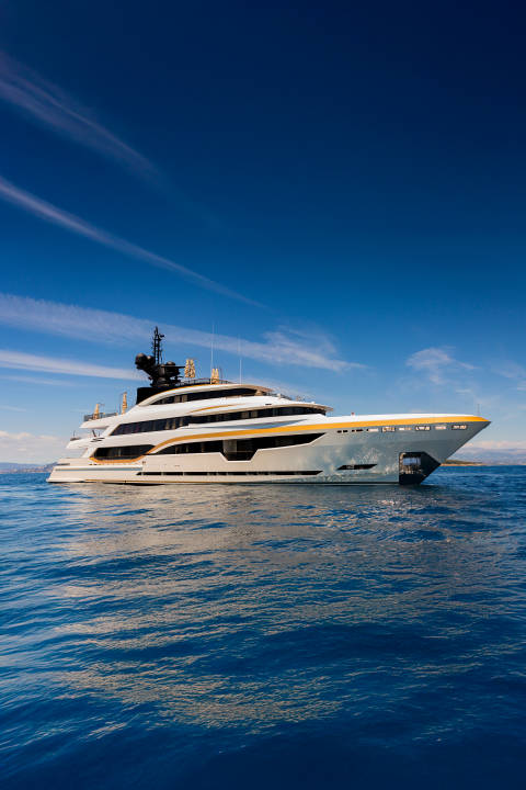 A luxurious white and gold yacht glides across calm blue water under a bright, cloud-streaked sky with distant green hills on the horizon.