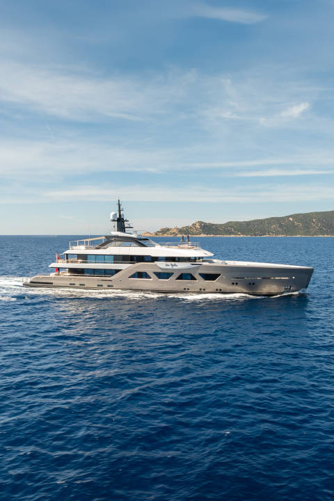 A sleek, modern yacht cruises through deep blue water with a hilly, tree-covered coastline in the background under a bright, cloudy sky.