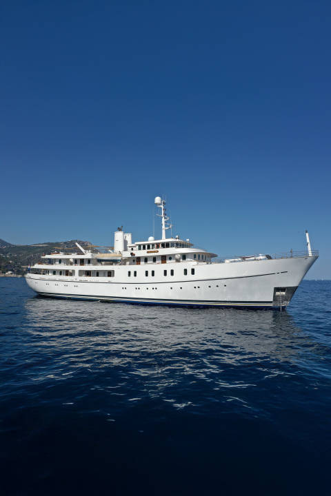 A large white yacht floats on calm blue water in the foreground, with a lush green coastline under a clear blue sky in the background.