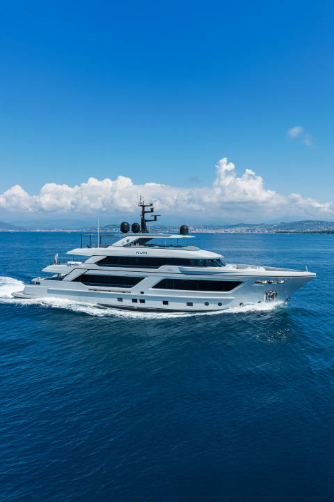 A sleek white yacht cruises through deep blue water with a hazy coastline and bright blue sky in the background.