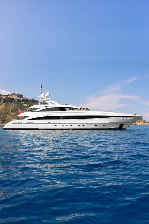 A sleek white yacht floats on the deep blue sea, with a lush green hillside dotted with buildings in the background under a clear sky.
