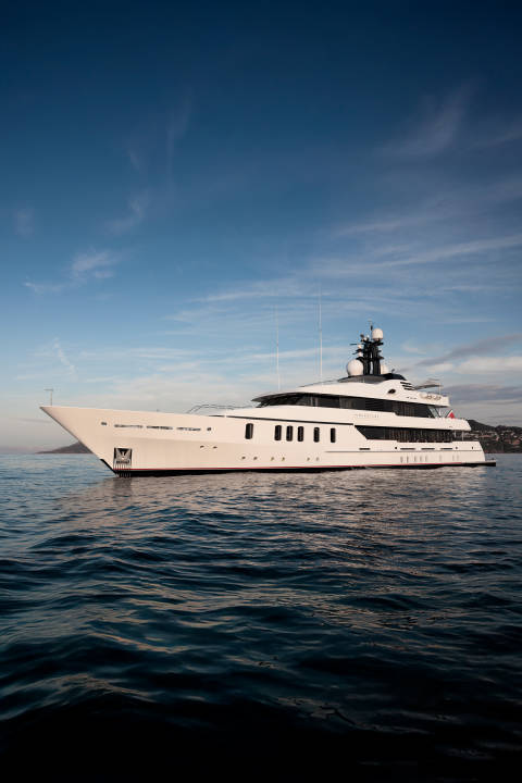 A large white yacht floats on calm blue water in the foreground, with a hazy coastline and a clear blue sky in the background.