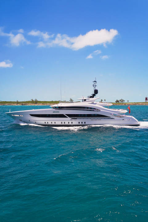 A sleek white yacht cruises through turquoise waters with a sandy coastline and blue sky in the background.