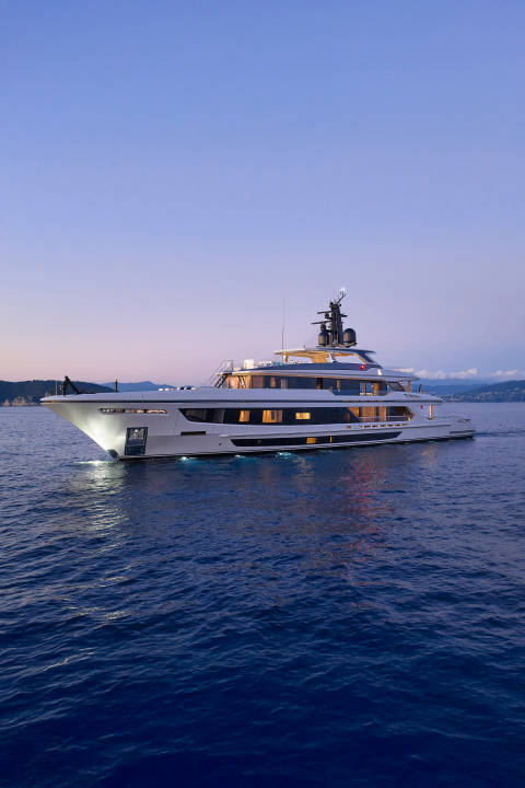 A large, illuminated yacht cruises on calm, dark blue water in the foreground, with a soft, twilight sky and distant, hazy landforms in the background.