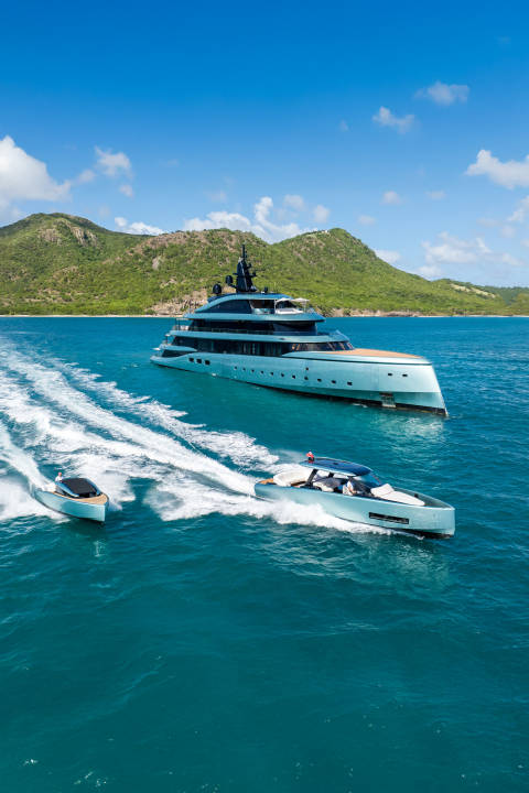 A large, sleek yacht is anchored in turquoise waters with two smaller speedboats zipping by in the foreground, all set against a backdrop of lush green hills under a clear blue sky.