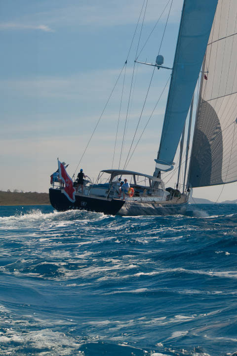 A sailboat cuts through choppy blue water in the foreground, with a distant, hazy coastline visible under a clear sky in the background.