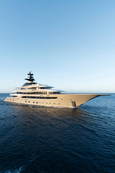 A large, luxurious yacht cruises on the deep blue ocean under a clear, pale blue sky.
