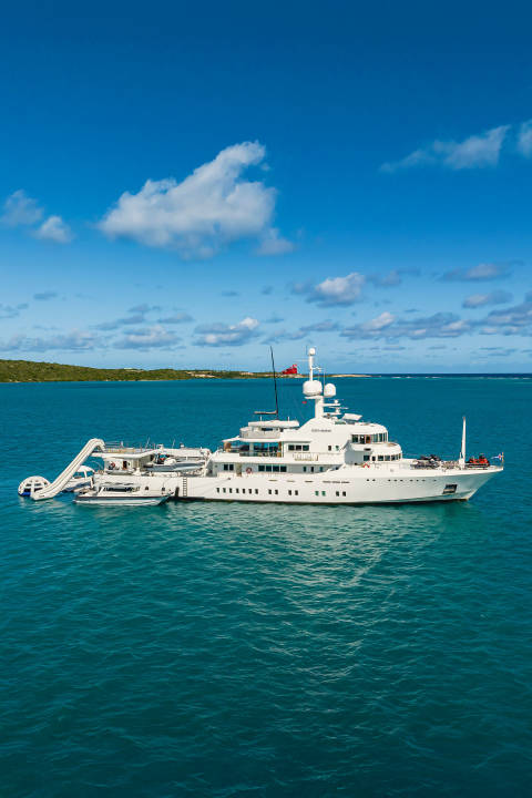 A large white yacht with a slide is anchored in turquoise water with a lush green island in the background under a blue sky with white clouds.