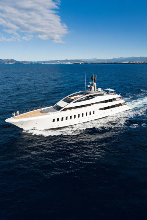 A sleek white yacht cruises through deep blue water, with a hazy coastline visible on the horizon under a clear sky.