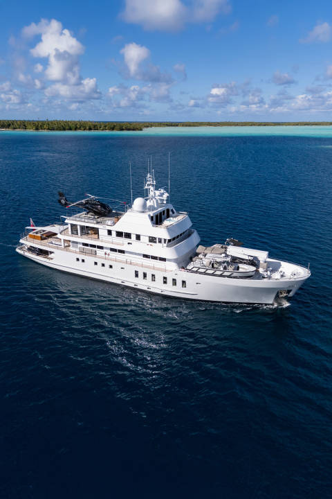 A large white yacht with a helicopter on its deck cruises through deep blue water, with a lush green island and a bright blue sky in the background.