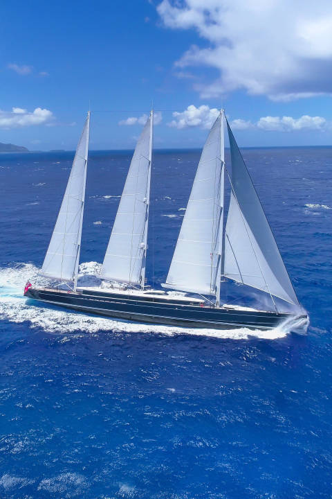 A majestic three-masted schooner glides through the deep blue ocean under a bright, cloudy sky.