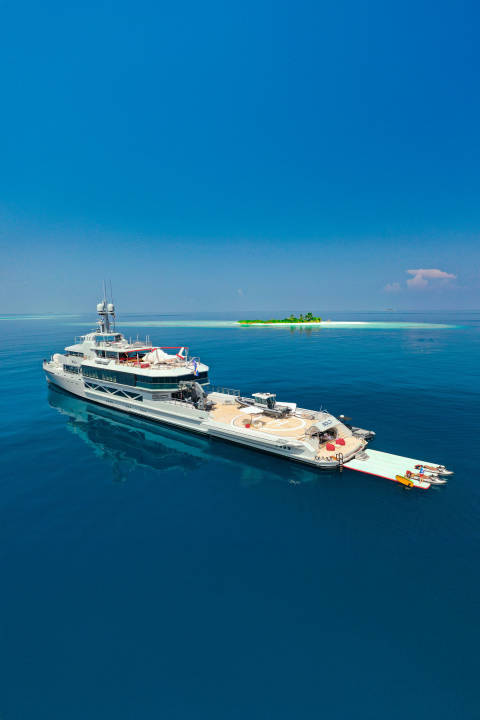 A large white yacht floats on calm blue water in the foreground, with a small green island visible in the distance under a clear blue sky.