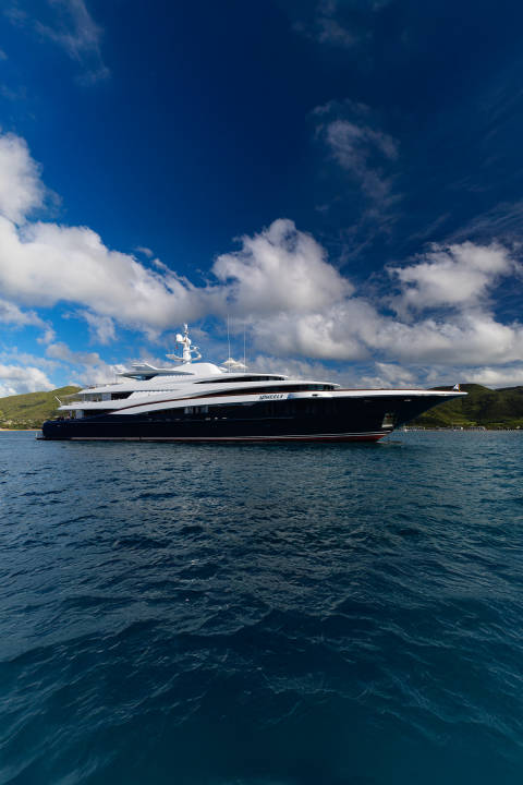 A large, dark yacht sails on calm blue water in the foreground, with lush green hills under a bright blue sky with white clouds in the background.
