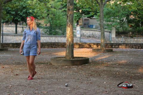 A woman in a denim romper stands in a park with petanque balls scattered on the ground, while trees and a stone wall form the background.