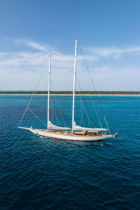 A majestic white sailboat glides across the deep blue ocean, with a lush green coastline and a clear blue sky in the background.
