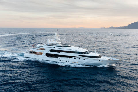 A large white yacht cruises through the deep blue ocean, with a hazy coastline visible in the distance under a soft sky.