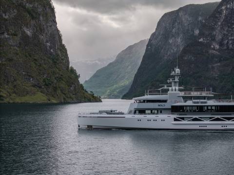 A sleek, modern yacht sails on calm water between towering, verdant cliffs under a cloudy sky.