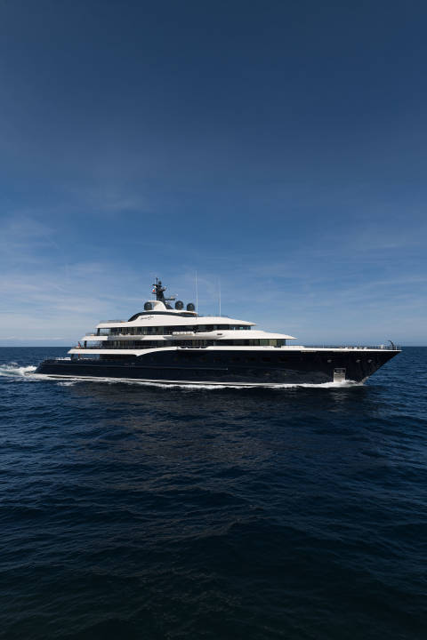 A large, dark blue yacht cruises on a calm, deep blue ocean under a clear, bright blue sky.
